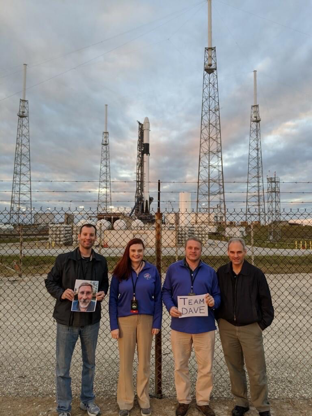 A few members of the Robotic Tool Stowage, or RiTS, team hold a sign and photo to represent Parker near a launch pad at Kennedy Space Center.